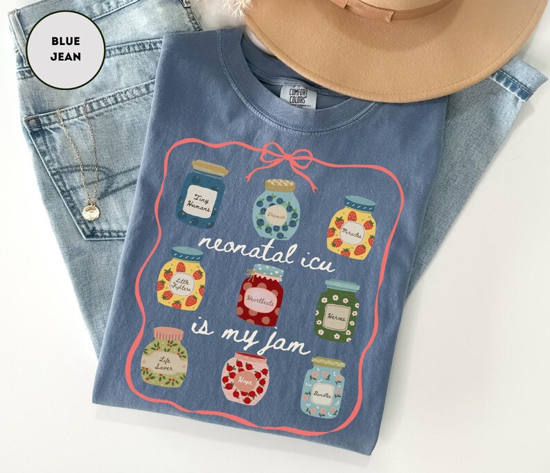 May include: A blue t-shirt with a graphic of jam jars and the text "neonatal icu is my jam." The shirt is laid on top of blue jeans and a tan hat. A gold necklace is also visible.