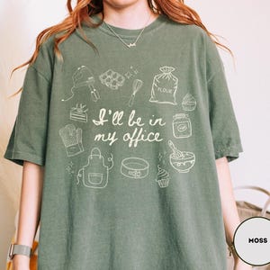 May include: Moss green t-shirt with a white graphic of baking tools and the text "I'll be in my office." The design includes a mixer, whisk, oven mitt, apron, cake pan, and cupcakes. The shirt is a comfortable, casual style.