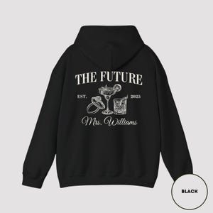 Pode incluir: Camisola de moletom com capuz preta com texto branco que diz "The Future Est. 2025 Mrs. Williams". O texto &eacute; cercado por uma ilustra&ccedil;&atilde;o de dois an&eacute;is e dois coquet&eacute;is.