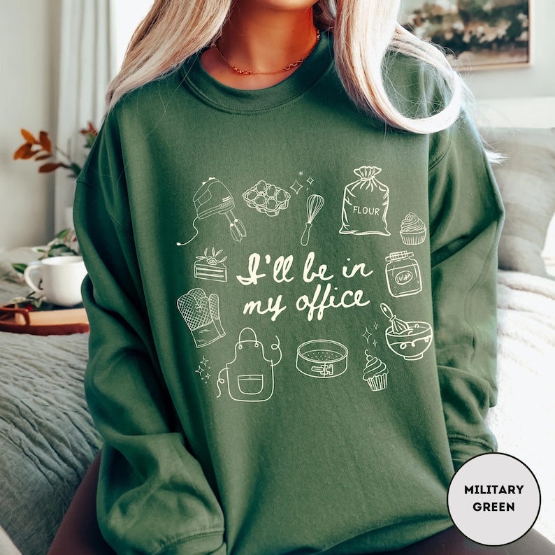 May include: A dark green sweatshirt with the text "I'll be in my office" and white illustrations of baking tools. The sweatshirt is a solid color with a crew neck and long sleeves. The color is described as "Military Green".