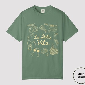 May include: Light green t-shirt with a cream-colored graphic design. The design includes illustrations of food items, wine glasses, and the text "La Dolce Vita". The shirt is a solid color and has a crew neck.
