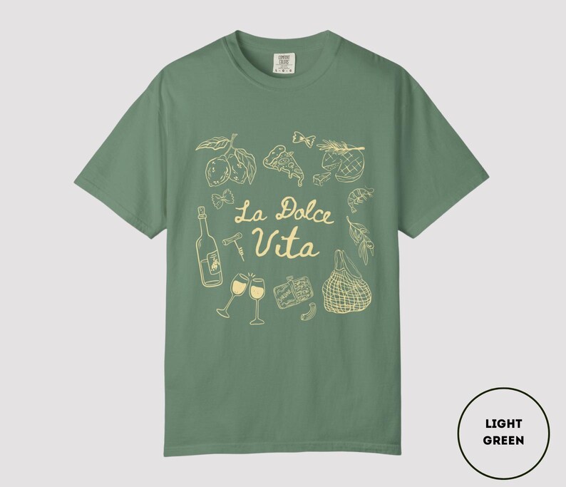 May include: Light green t-shirt with a cream-colored graphic design. The design includes illustrations of food items, wine glasses, and the text "La Dolce Vita". The shirt is a solid color and has a crew neck.