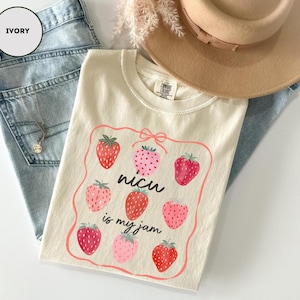 May include: Ivory-colored t-shirt with a strawberry graphic and the text "nicu is my jam". The shirt is folded on top of light blue denim jeans. A tan hat and a gold necklace are also visible.