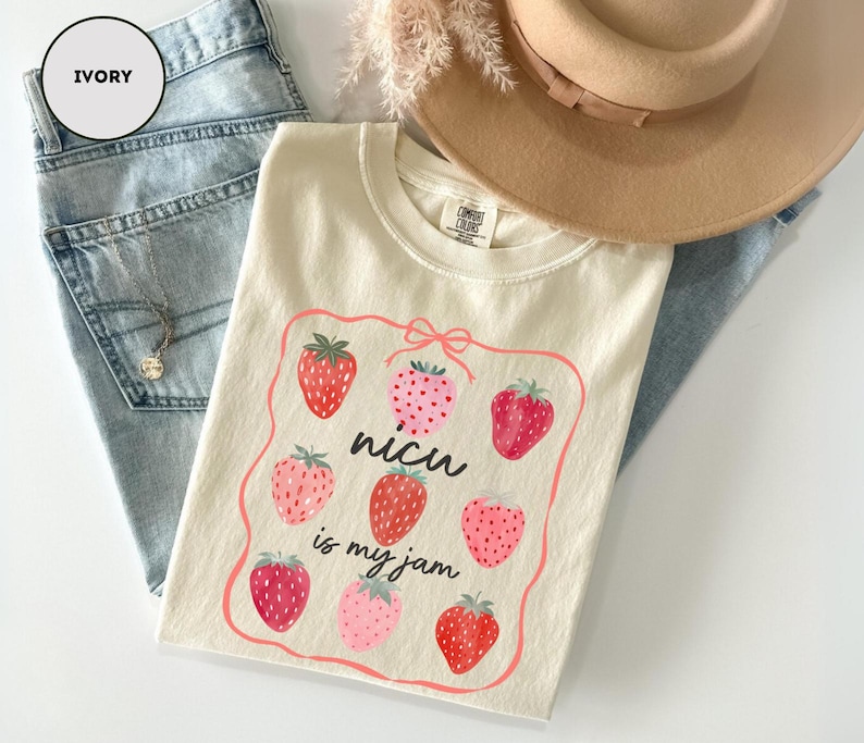 May include: Ivory-colored t-shirt with a strawberry graphic and the text "nicu is my jam". The shirt is folded on top of light blue denim jeans. A tan hat and a gold necklace are also visible.