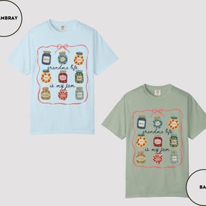 Peut inclure: Deux t-shirts &agrave; manches courtes, un bleu clair et un vert sauge. Chaque t-shirt pr&eacute;sente un motif de bocaux de confiture avec le texte "grandma life is my jam" dans un cadre rose.