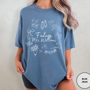 May include: A blue jean-colored t-shirt with white line art illustrations of wedding-related icons. The text "Future Mrs. Williams" is centered on the shirt. The design includes champagne glasses, shoes, doves, and a banner that says "Bride".