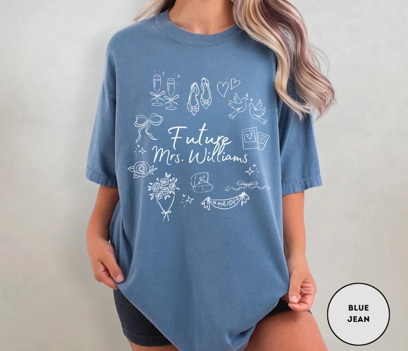 May include: A blue jean-colored t-shirt with white line art illustrations of wedding-related icons. The text "Future Mrs. Williams" is centered on the shirt. The design includes champagne glasses, shoes, doves, and a banner that says "Bride".