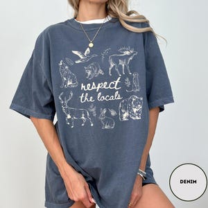 May include: Denim blue t-shirt with a white graphic of various animals and the text "respect the locals". The design includes a wolf, bear, deer, and other wildlife. The shirt has short sleeves and a relaxed fit.