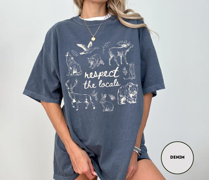 May include: Denim blue t-shirt with a white graphic of various animals and the text "respect the locals". The design includes a wolf, bear, deer, and other wildlife. The shirt has short sleeves and a relaxed fit.