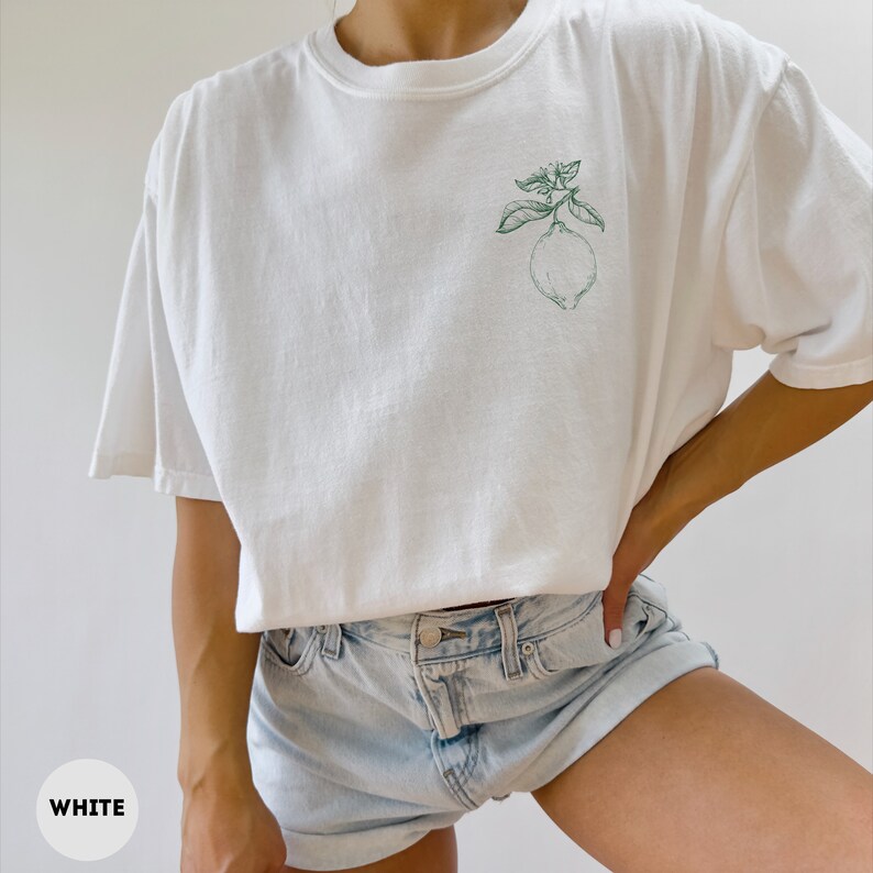 May include: White t-shirt with a green lemon and leaf design on the front. The shirt is a relaxed fit and has short sleeves. The word "WHITE" is in a white circle in the bottom left corner.