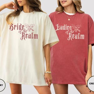 May include: Two oversized t-shirts, one ivory and one crimson, featuring the text "Bride of the Realm" and "Ladies of the Realm" in a decorative font. The shirts have short sleeves and a relaxed fit, perfect for casual wear.