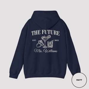 Pode incluir: Camisola de capuz azul marinho com texto branco que diz "The Future Est. 2025 Mrs. Williams" com um gr&aacute;fico de dois copos de cocktail e dois an&eacute;is.