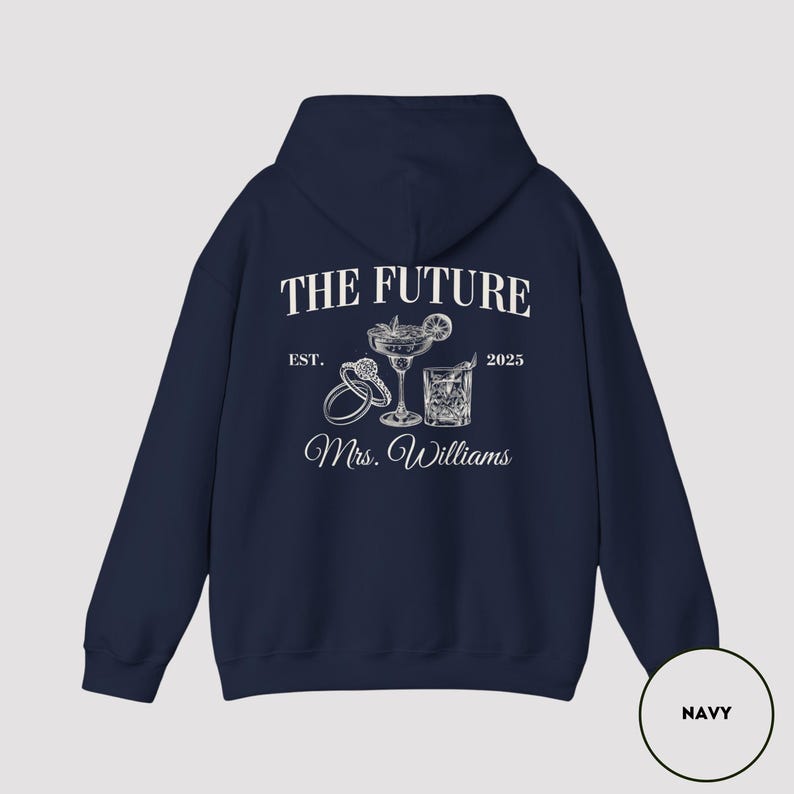 Pode incluir: Camisola de capuz azul marinho com texto branco que diz "The Future Est. 2025 Mrs. Williams" com um gr&aacute;fico de dois copos de cocktail e dois an&eacute;is.