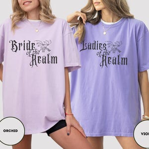 May include: Two oversized t-shirts, one orchid and one violet, with the text "Bride of the Realm" and "Ladies of the Realm" in a gothic font. The shirts have a relaxed fit and short sleeves. The image includes color swatches labeled "Orchid" and "Violet".