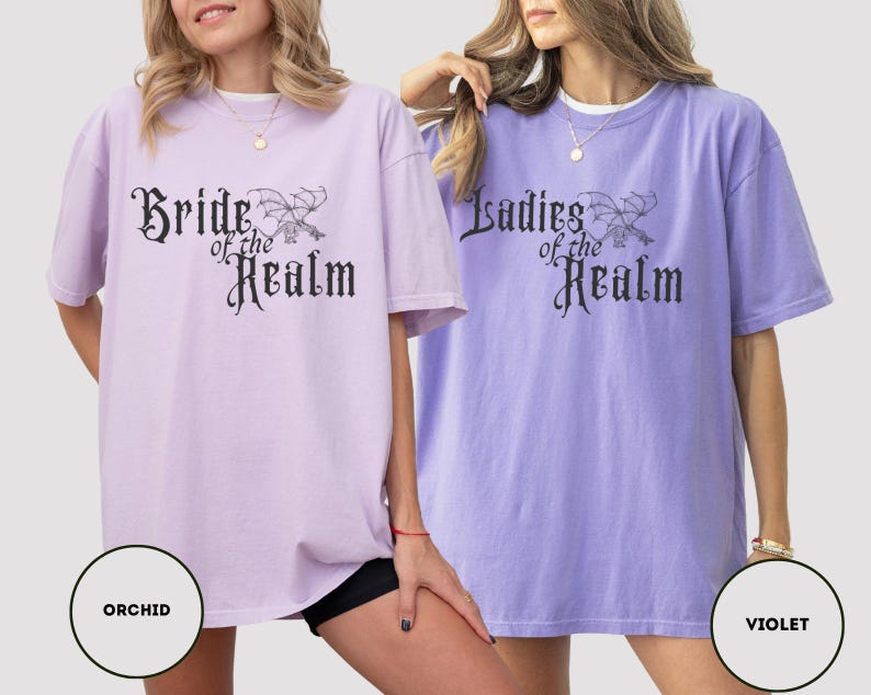 May include: Two oversized t-shirts, one orchid and one violet, with the text "Bride of the Realm" and "Ladies of the Realm" in a gothic font. The shirts have a relaxed fit and short sleeves. The image includes color swatches labeled "Orchid" and "Violet".