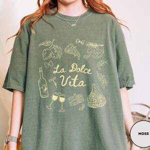 May include: Moss green t-shirt with a yellow graphic design. The design includes illustrations of food items like lemons, pizza, and wine glasses, along with the text "La Dolce Vita". The shirt has short sleeves and a relaxed fit.