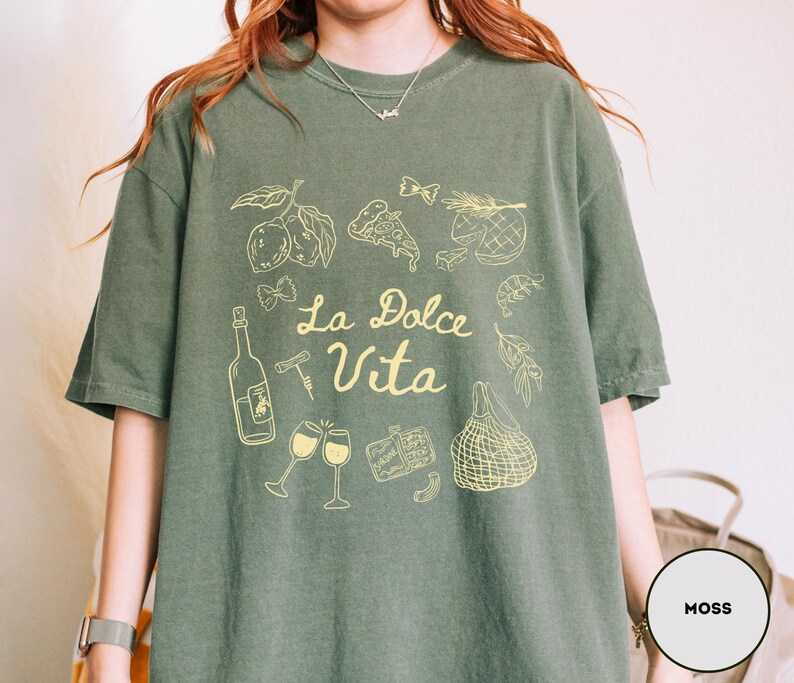 May include: Moss green t-shirt with a yellow graphic design. The design includes illustrations of food items like lemons, pizza, and wine glasses, along with the text "La Dolce Vita". The shirt has short sleeves and a relaxed fit.