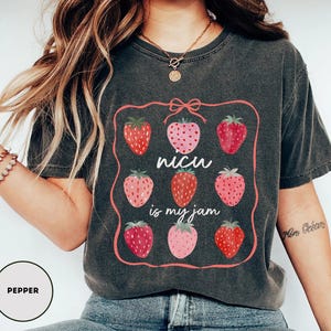 May include: Dark grey t-shirt featuring a pink framed design with nine strawberries in varying shades of red and pink. The text "NICU is my jam" is in the center. The shirt is a casual style.