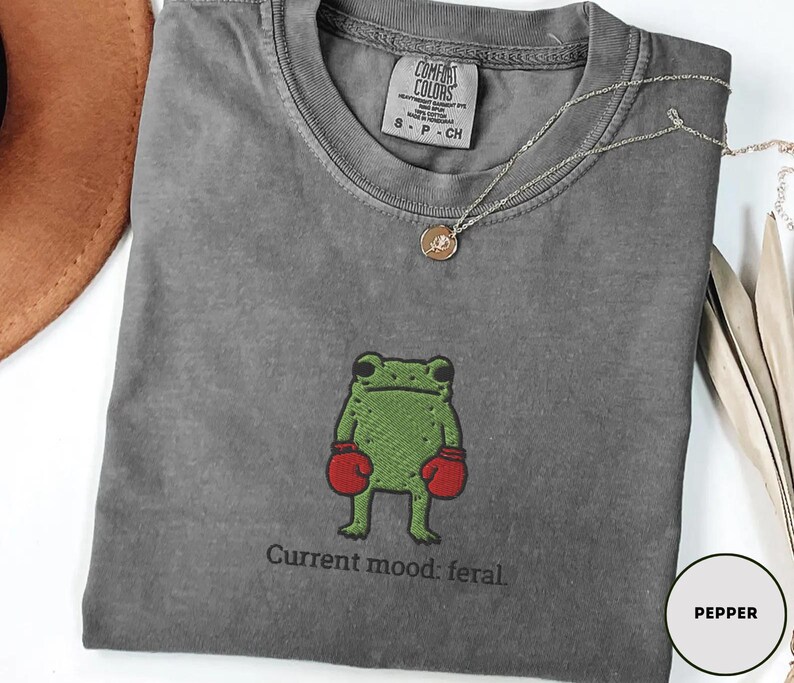 May include: A gray t-shirt with a green frog wearing red boxing gloves. The text "Current mood: feral" is below the frog. A silver necklace with a circular pendant is draped on the shirt.