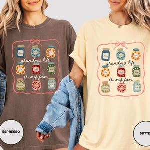 Peut inclure: Deux t-shirts, un espresso et un beurre, pr&eacute;sentent un graphisme color&eacute; de pots de confiture dans un cadre rose. Le texte "grandma life is my jam" est centr&eacute; en dessous. Les t-shirts sont d&eacute;contract&eacute;s.