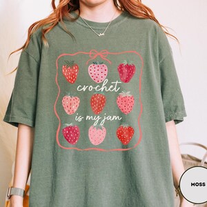 Puede incluir: Camiseta verde musgo con un gr&aacute;fico de fresas y el texto "crochet is my jam". El gr&aacute;fico presenta un marco rosa con nueve fresas en varios tonos de rojo y rosa.