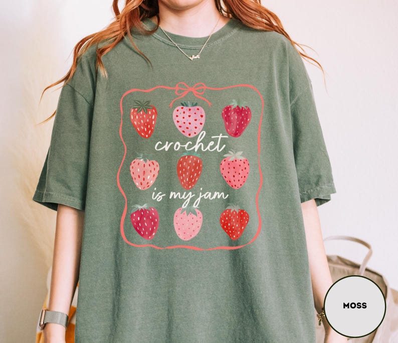 Puede incluir: Camiseta verde musgo con un gr&aacute;fico de fresas y el texto "crochet is my jam". El gr&aacute;fico presenta un marco rosa con nueve fresas en varios tonos de rojo y rosa.