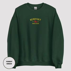 May include: Forest green sweatshirt featuring the word "NEWPORT" in yellow arched text, with "16 39" and "RHODE ISLAND" in smaller text. A red lobster graphic is below the text. The sweatshirt is a Gildan brand.