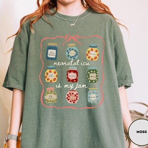 May include: Moss green t-shirt featuring a graphic of nine jam jars with different fruit designs. The text "neonatal icu is my jam" is written in a pink frame. The shirt is made of a soft material.