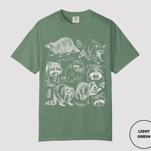 Op de afbeelding: Lichtgroen T-shirt met een witte illustratie van wasberen. Het ontwerp bevat gedetailleerde tekeningen van wasberen in verschillende poses en close-up gezichten. Het T-shirt heeft een effen lichtgroene kleur.