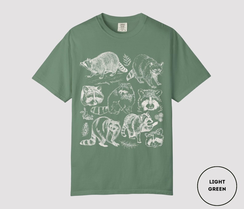 Op de afbeelding: Lichtgroen T-shirt met een witte illustratie van wasberen. Het ontwerp bevat gedetailleerde tekeningen van wasberen in verschillende poses en close-up gezichten. Het T-shirt heeft een effen lichtgroene kleur.