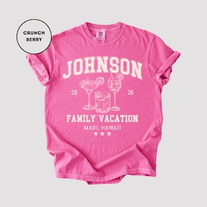 K&ouml;nnte beinhalten: Ein pinkfarbenes T-Shirt mit hochgekrempelten &Auml;rmeln. Das Shirt zeigt den Namen "JOHNSON" in wei&szlig;en Blockbuchstaben &uuml;ber einer Grafik von drei Cocktailgl&auml;sern und dem Text "FAMILY VACATION MAUI, HAWAII". Das Shirt ist mit "Crunch Berry" beschriftet.