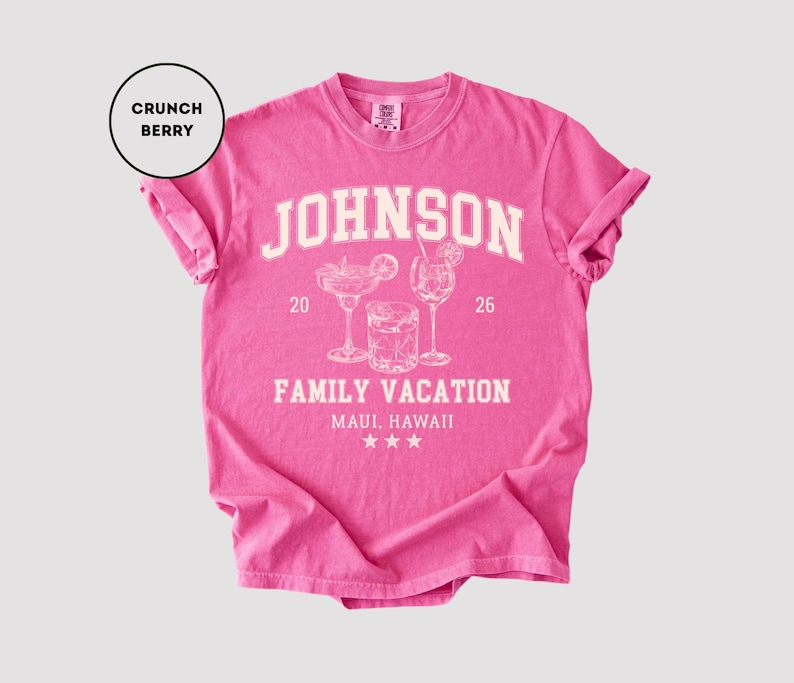 K&ouml;nnte beinhalten: Ein pinkfarbenes T-Shirt mit hochgekrempelten &Auml;rmeln. Das Shirt zeigt den Namen "JOHNSON" in wei&szlig;en Blockbuchstaben &uuml;ber einer Grafik von drei Cocktailgl&auml;sern und dem Text "FAMILY VACATION MAUI, HAWAII". Das Shirt ist mit "Crunch Berry" beschriftet.