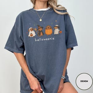 Op de afbeelding: Een denimblauw t-shirt met een afbeelding van vier teckels in Halloween-kostuums, waaronder een spook, een heks, een pompoen en een mummie. De tekst "halloweenie" staat onder de honden gedrukt, vergezeld door kleine sterpictogrammen.