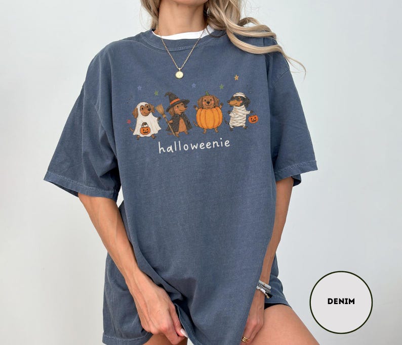 Op de afbeelding: Een denimblauw t-shirt met een afbeelding van vier teckels in Halloween-kostuums, waaronder een spook, een heks, een pompoen en een mummie. De tekst "halloweenie" staat onder de honden gedrukt, vergezeld door kleine sterpictogrammen.