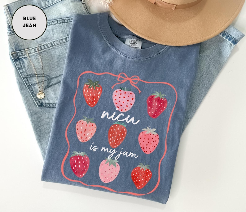 May include: A blue t-shirt with a strawberry graphic and the text "nicu is my jam". The shirt is folded on top of a pair of blue jeans. A tan hat is in the background.
