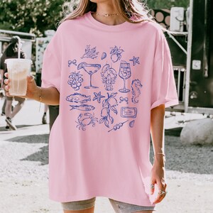 May include: A light pink t-shirt featuring a blue illustration of seafood and cocktails. The design includes images of fish, a lobster, a seahorse, and a martini glass. The word "Sardines" is also visible in the design.