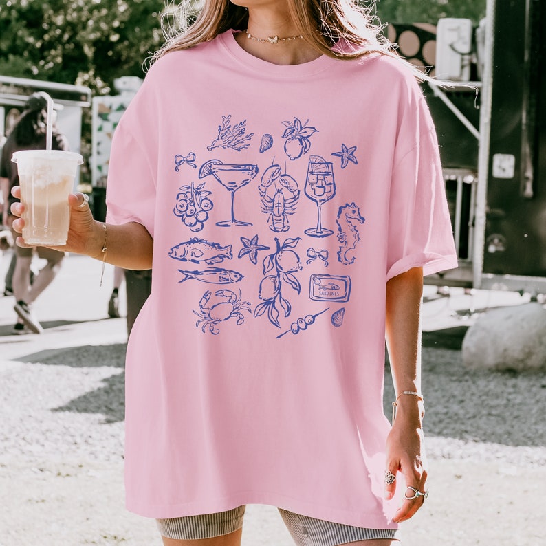 May include: A light pink t-shirt featuring a blue illustration of seafood and cocktails. The design includes images of fish, a lobster, a seahorse, and a martini glass. The word "Sardines" is also visible in the design.