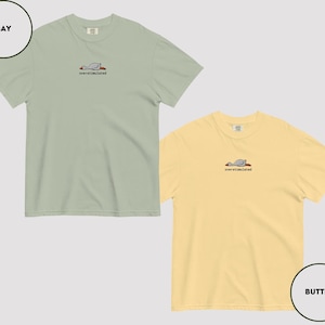 May include: Two short-sleeved t-shirts, one in a bay colour and the other in a butter colour, are shown. Each shirt has a small embroidered design of a resting bird with the word "overstimulated" beneath. These are casual wear items.