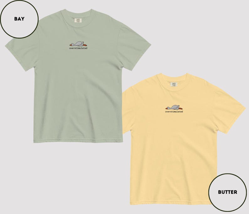 May include: Two short-sleeved t-shirts, one in a bay colour and the other in a butter colour, are shown. Each shirt has a small embroidered design of a resting bird with the word "overstimulated" beneath. These are casual wear items.