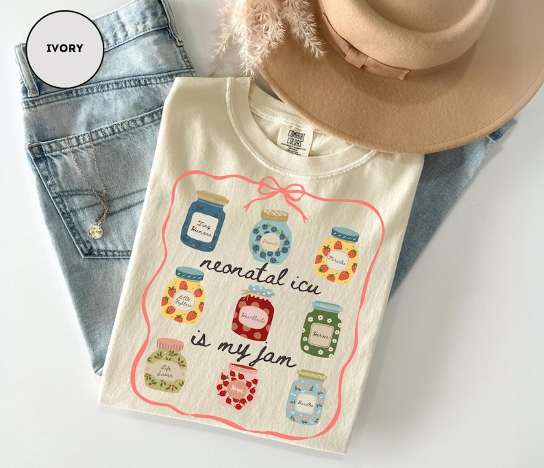 May include: Ivory-colored t-shirt with a colorful graphic of jam jars and the text "neonatal ICU is my jam." The shirt is laid on top of light blue jeans, a necklace, and a tan hat.
