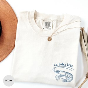 Puede incluir: Camiseta de color marfil con un gr&aacute;fico de camar&oacute;n azul y el texto "La Dolce Vita" impreso en la parte delantera.