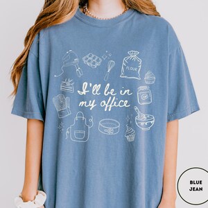 May include: Blue jean-colored t-shirt with a white graphic of baking tools and the text "I'll be in my office." The design includes a mixer, whisk, oven mitt, apron, cake pan, and cupcakes. The shirt is a comfortable, casual style.