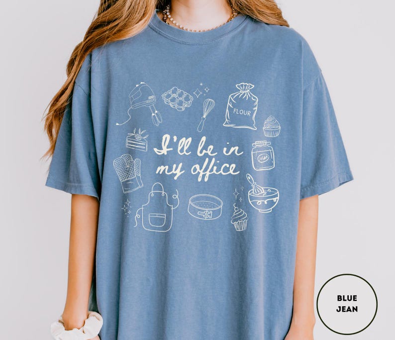 May include: Blue jean-colored t-shirt with a white graphic of baking tools and the text "I'll be in my office." The design includes a mixer, whisk, oven mitt, apron, cake pan, and cupcakes. The shirt is a comfortable, casual style.