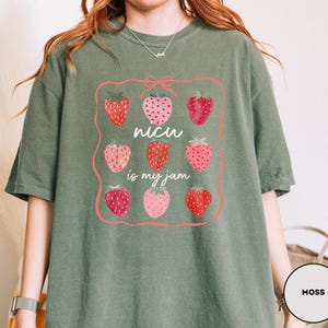 May include: Moss green t-shirt with a strawberry graphic. The graphic features nine strawberries in a pink frame, with the text "NICU is my jam" in white. The shirt is made of a soft material.