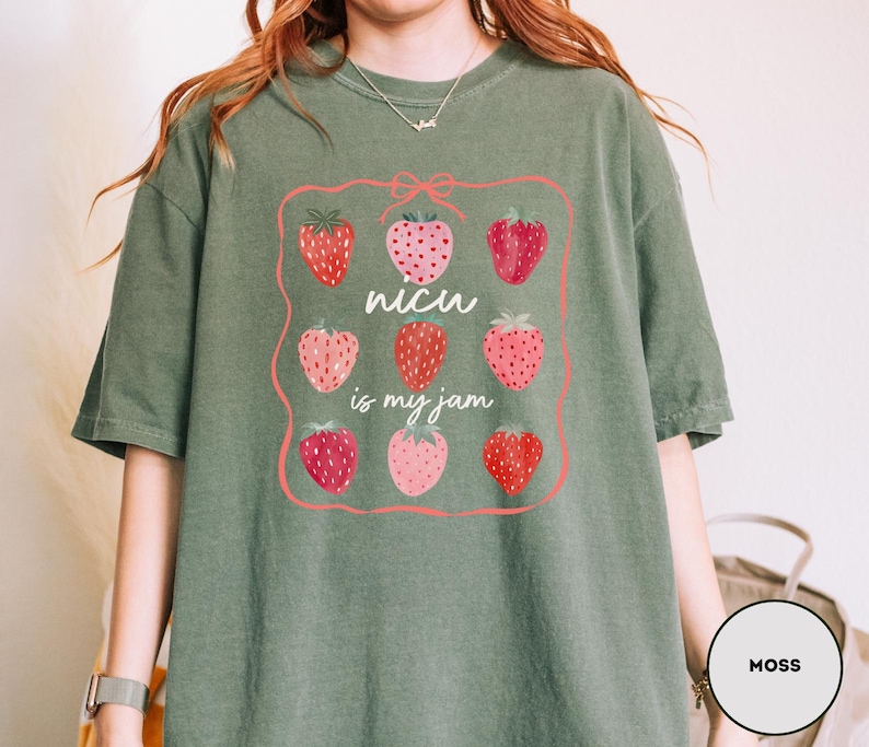 May include: Moss green t-shirt with a strawberry graphic. The graphic features nine strawberries in a pink frame, with the text "NICU is my jam" in white. The shirt is made of a soft material.