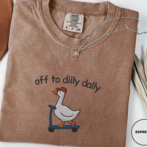 May include: Espresso-colored t-shirt with the phrase "off to dilly dally" above an embroidered cartoon goose wearing a red cap and riding a blue scooter. The shirt has a Comfort Colors tag and a silver necklace with a pendant.
