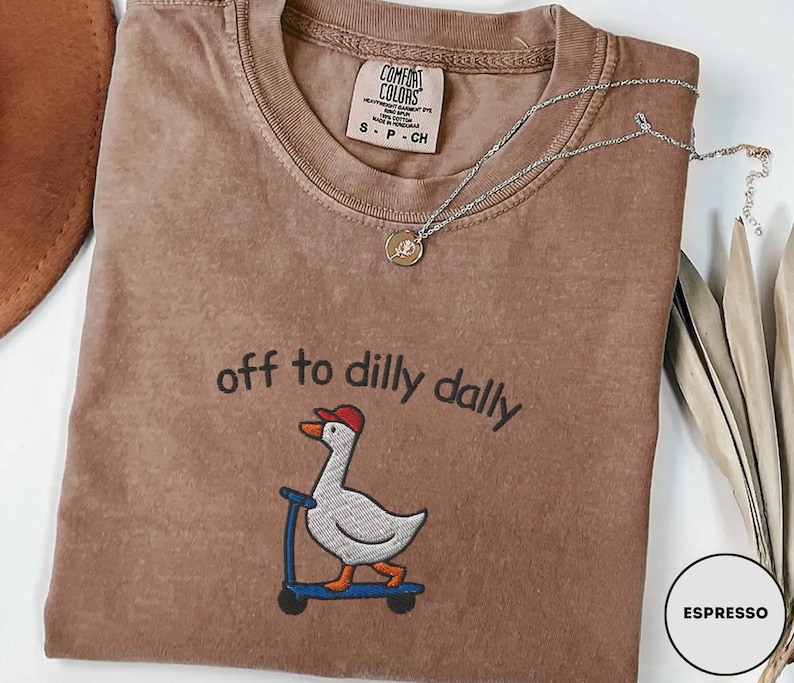 May include: Espresso-colored t-shirt with the phrase "off to dilly dally" above an embroidered cartoon goose wearing a red cap and riding a blue scooter. The shirt has a Comfort Colors tag and a silver necklace with a pendant.