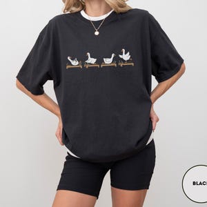 Puede incluir: Camiseta negra con cuello redondo blanco, con un dise&ntilde;o caprichoso de cuatro gansos blancos sobre plataformas de madera. Los gansos est&aacute;n en varias poses. La camiseta es de corte holgado.