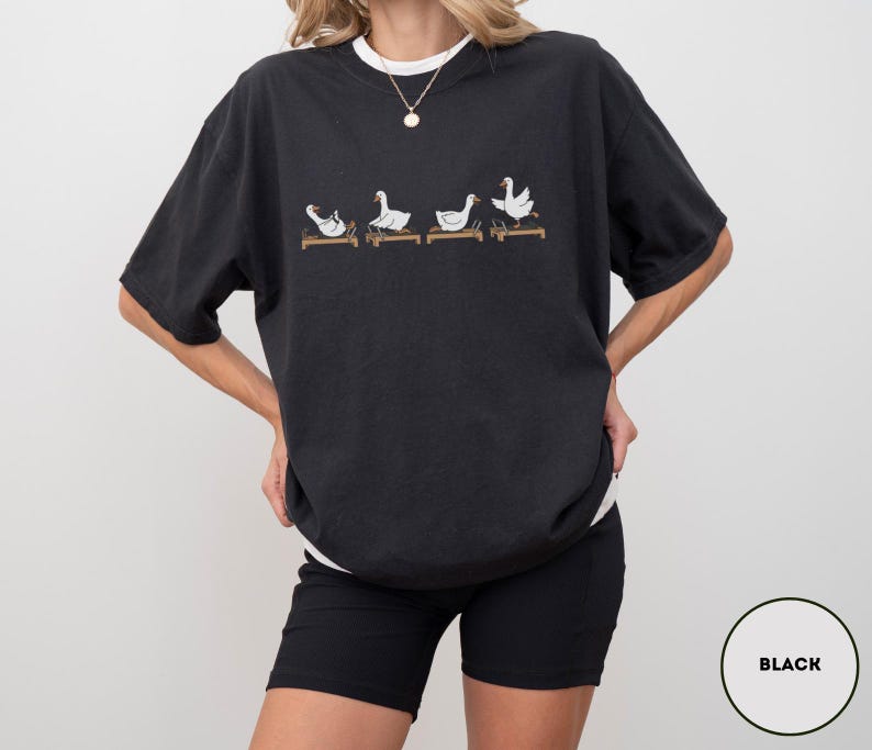 Puede incluir: Camiseta negra con cuello redondo blanco, con un dise&ntilde;o caprichoso de cuatro gansos blancos sobre plataformas de madera. Los gansos est&aacute;n en varias poses. La camiseta es de corte holgado.