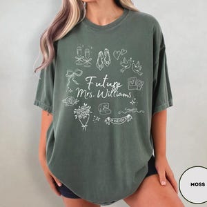 May include: Moss green t-shirt with a white graphic design. The design includes champagne glasses, shoes, doves, hearts, and the text "Future Mrs. Williams". A bouquet of flowers, a ring, and a banner that says "Bride" are also included.